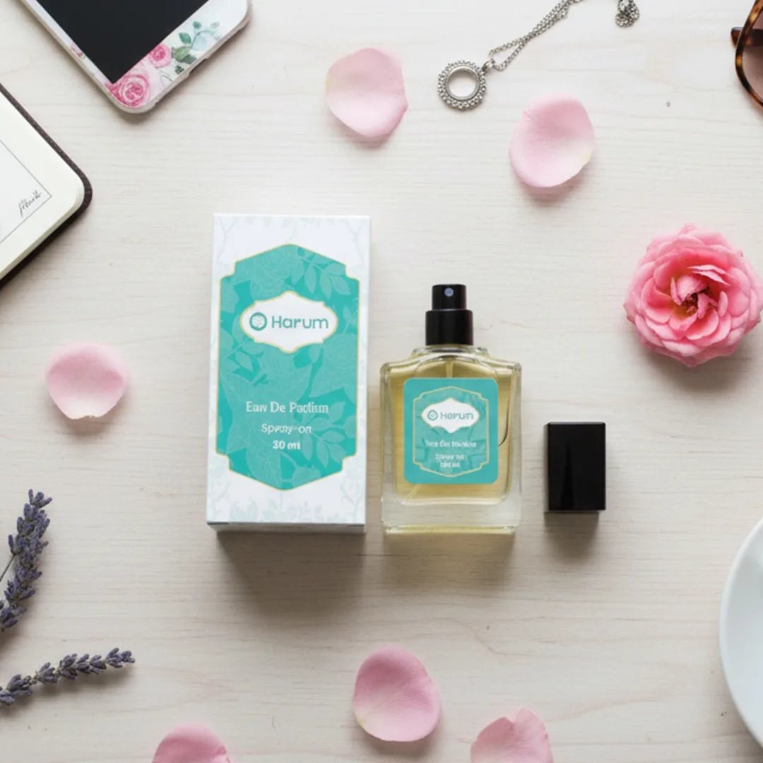 A flat lay of Harum.sg Eau De Parfum 30ml bottle and box on a light wooden desk, surrounded by rose petals, flowers, jewelry, and a smartphone for a feminine lifestyle aesthetic.