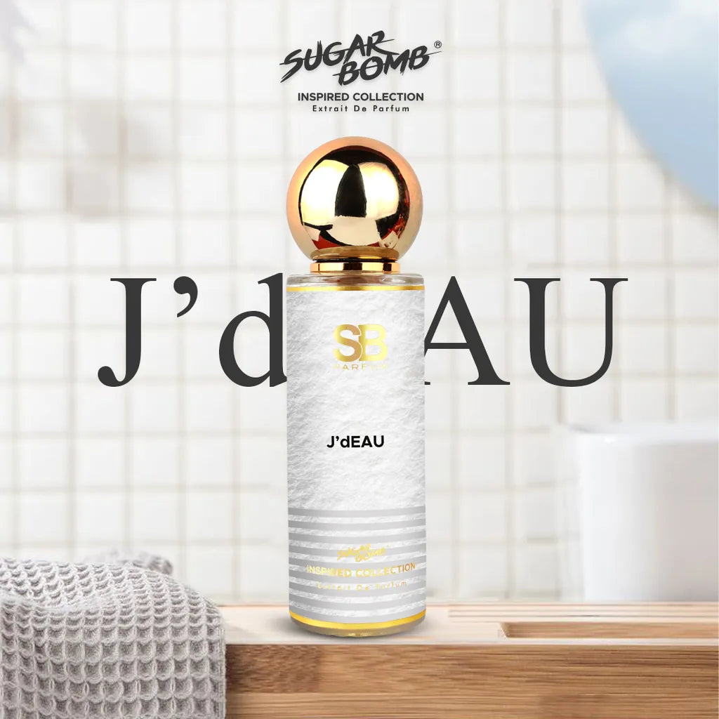 SugarBomb J'dEAU Extrait De Parfum bottle from the Inspired Collection, standing on a light wood counter next to a folded grey towel in a bright, tiled modern bathroom.