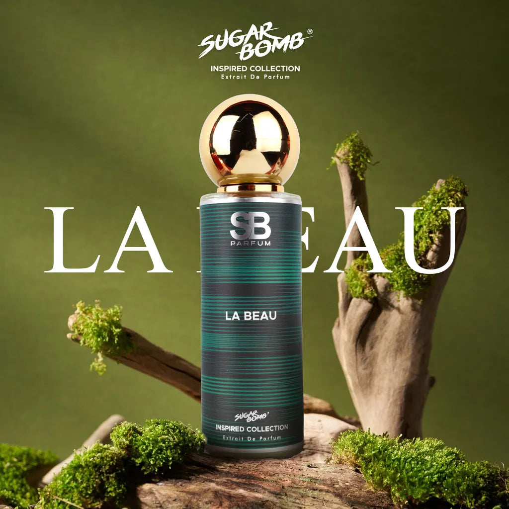 SugarBomb La Beau Extrait De Parfum bottle from the Inspired Collection, a clone fragrance inspired by Jean Paul Gaultier Le Beau. The product shot shows the green striped perfume bottle with a metallic gold ball cap centered on a rustic piece of wood covered in green moss against a soft green background.