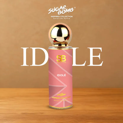SugarBomb Idole Extrait De Parfum bottle from the Inspired Collection, standing on a glassy pink platform covered in fresh morning dew droplets and scattered pink rose petals.
