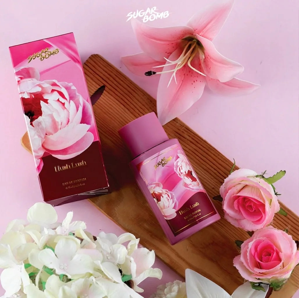 SugarBomb Hush Lush Eau De Parfum bottle and packaging displayed on a wooden board surrounded by pink lilies and roses.
